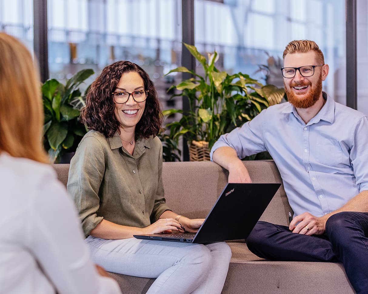 A man and woman sitting on a couch laughing. The woman has a laptop sitting on her lap.