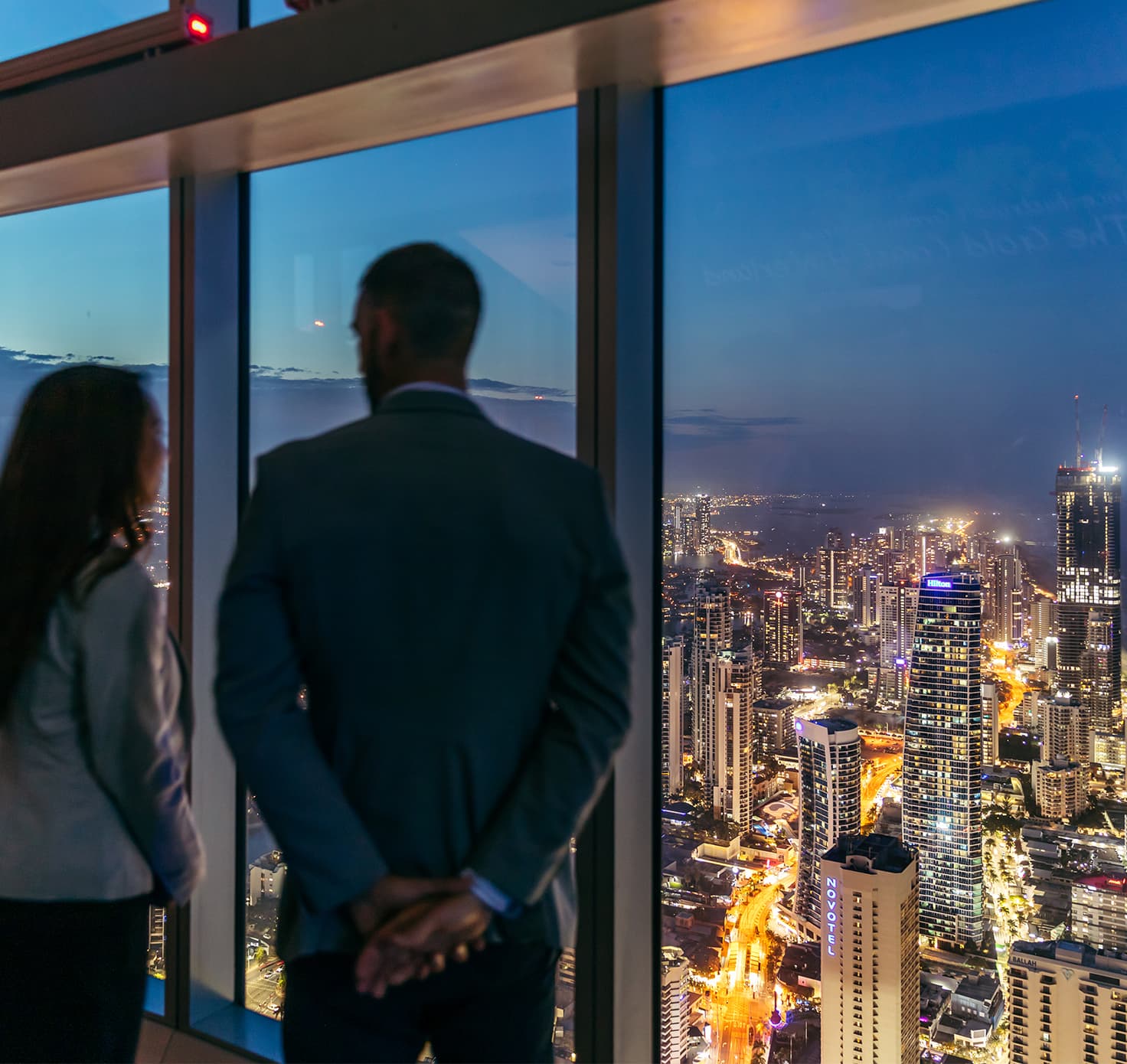 Two professional people look out the windows of the Q1 high-rise at Surfers Paradise below.