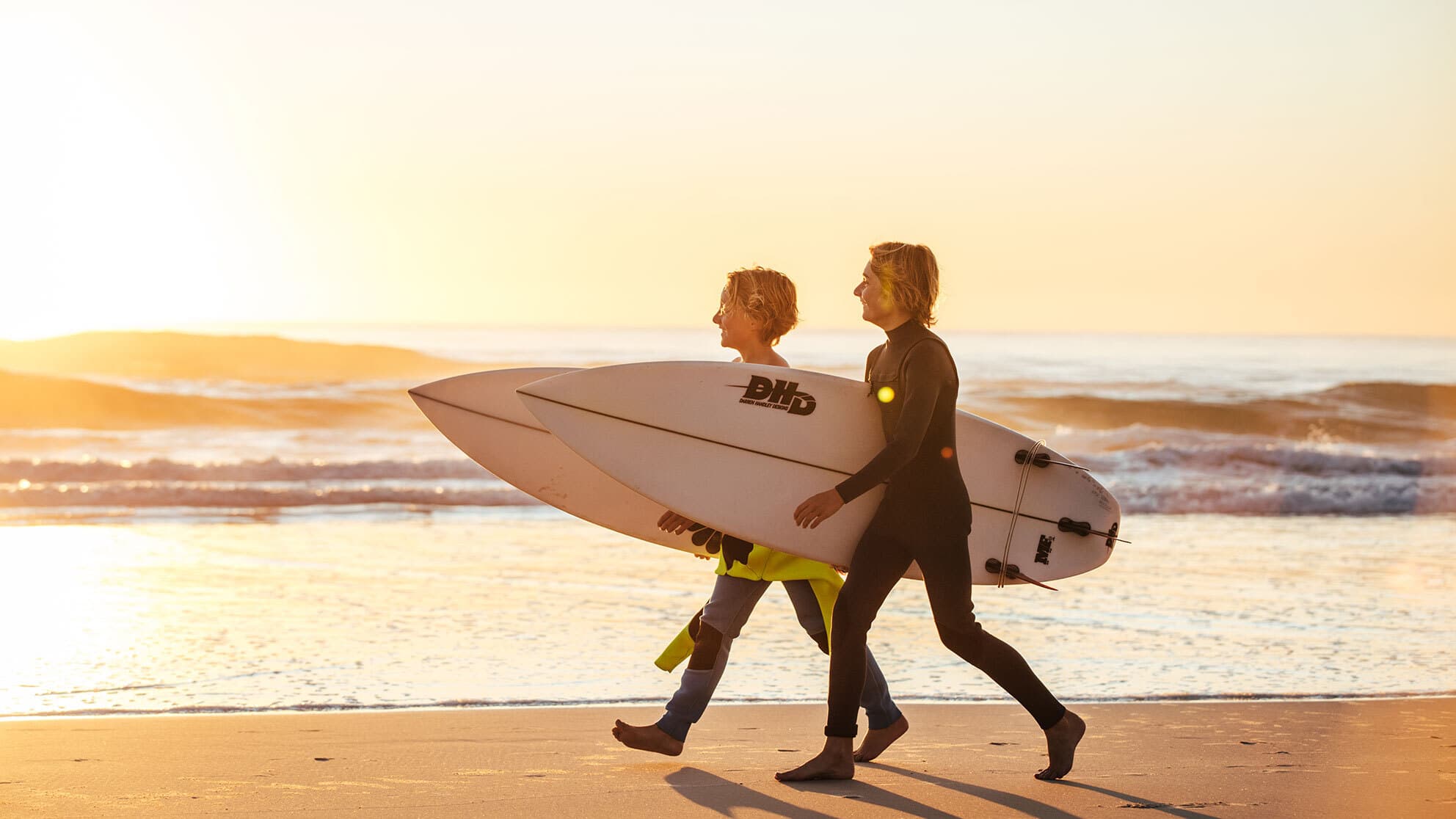 Two young boys walk along the beach carrying surfboards.